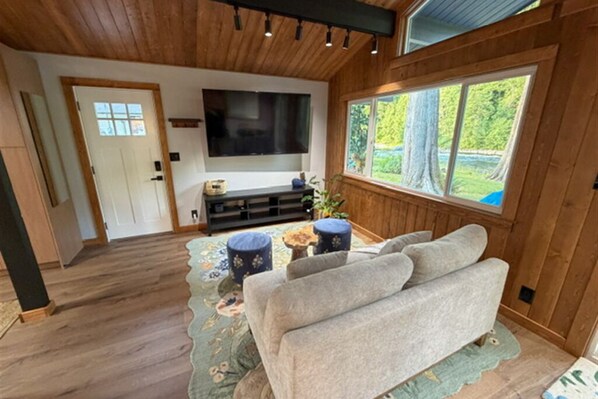 A cozy living room with a sofa, a flat-screen TV, and a window with a view of trees.