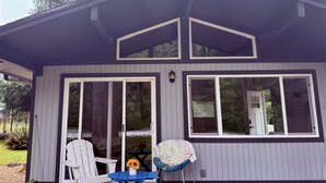 En enplansbyggnad med en mörk fasad och vita detaljer. Det finns två stora fönster på framsidan, varav det ena har en dörr och det andra inte. Utanför står en vit stol och ett blått bord med en solros på.