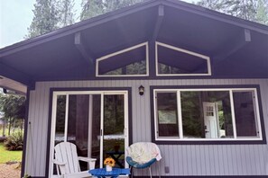 A single-story building with a dark exterior and white trim. There are two large windows on the front, one with a door and one without. Outside, there is a white chair and a blue table with a sunflower.