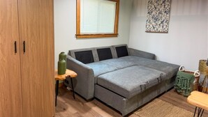 Ett kompakt vardagsrum med en grå soffa, en trägarderob, ett litet bord med en grön kruka och ett fönster med persienner.