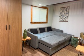A compact living space with a grey sofa, a wooden wardrobe, a small table with a green container, and a window with blinds.