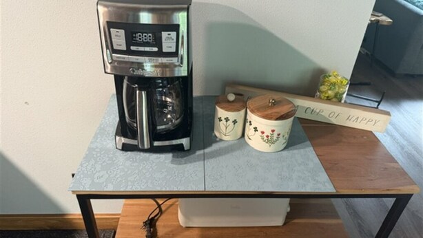 En kaffebryggare står på ett bord tillsammans med två dekorativa behållare och en skylt med texten ”En kopp lycka”.