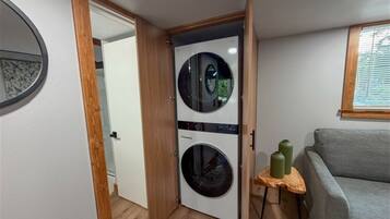 Een wasserette met een voorlader wasmachine en droger, een klein tafeltje met twee groene flessen, en een grijze bank in de aangrenzende woonruimte.