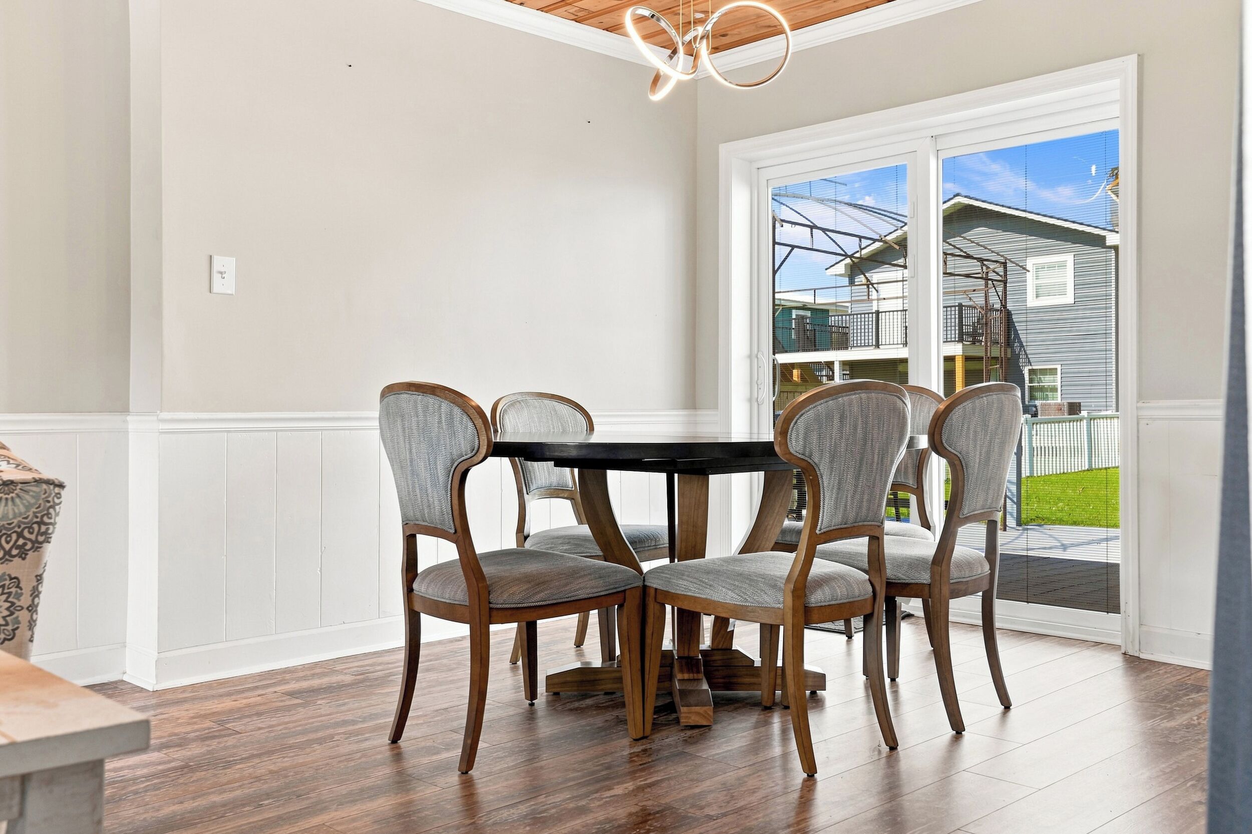 Un comedor con una mesa de madera y cuatro sillas, una pared blanca y un ventanal con vista a una casa y una valla.