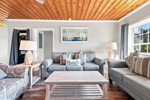 A living room with a wooden ceiling, a grey sofa, a coffee table, and a painting of sailboats. - Sea La Vie | Coastal Comfort by the Beach (North Myrtle Beach)