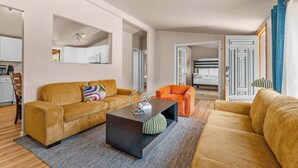 Uma sala moderna com sofá, poltrona e mesa de centro, uma cozinha com armários e micro-ondas, e um quarto com cama e cortinas.