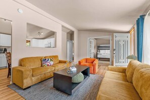 Ein modernes Wohnzimmer mit Sofa, Sessel und Couchtisch, ein Küchenbereich mit Schränken und einer Mikrowelle sowie ein Schlafzimmer mit Bett und Vorhängen.