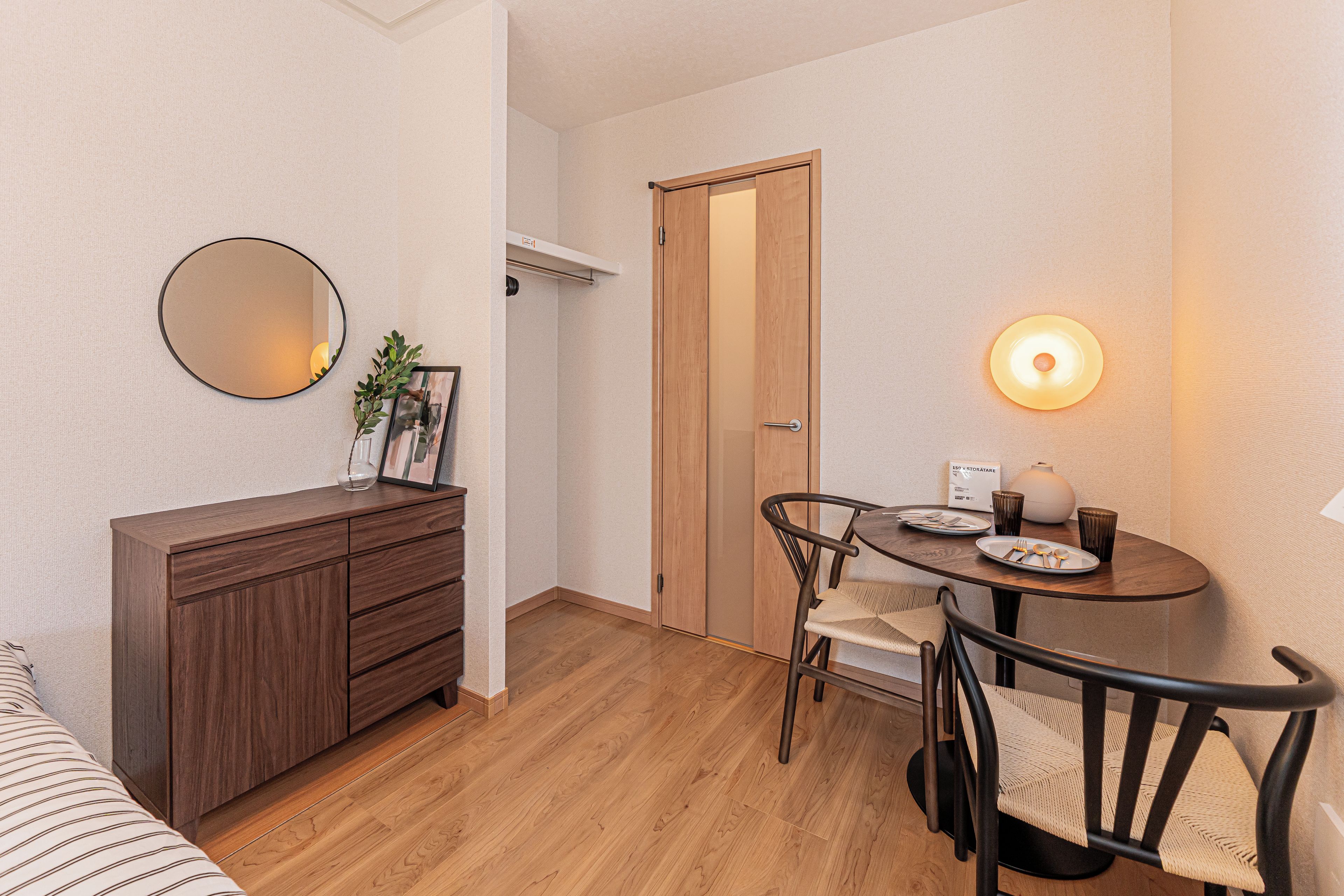 A compact room with a wooden cabinet, a round table with chairs, a mirror, and a wall-mounted light fixture.