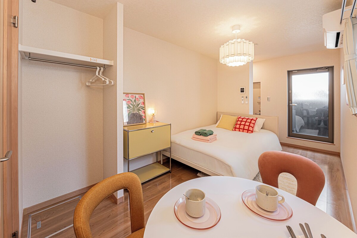 A hotel room with a bed, bedside table, a hanging light fixture, a window, and a dining area with a table and chairs.