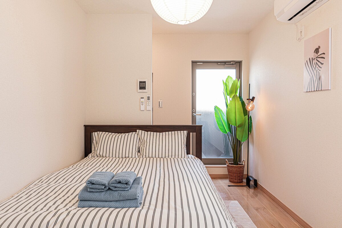 A hotel room with a bed, a potted plant, a wall-mounted air conditioning unit, and a door with a frosted glass window.