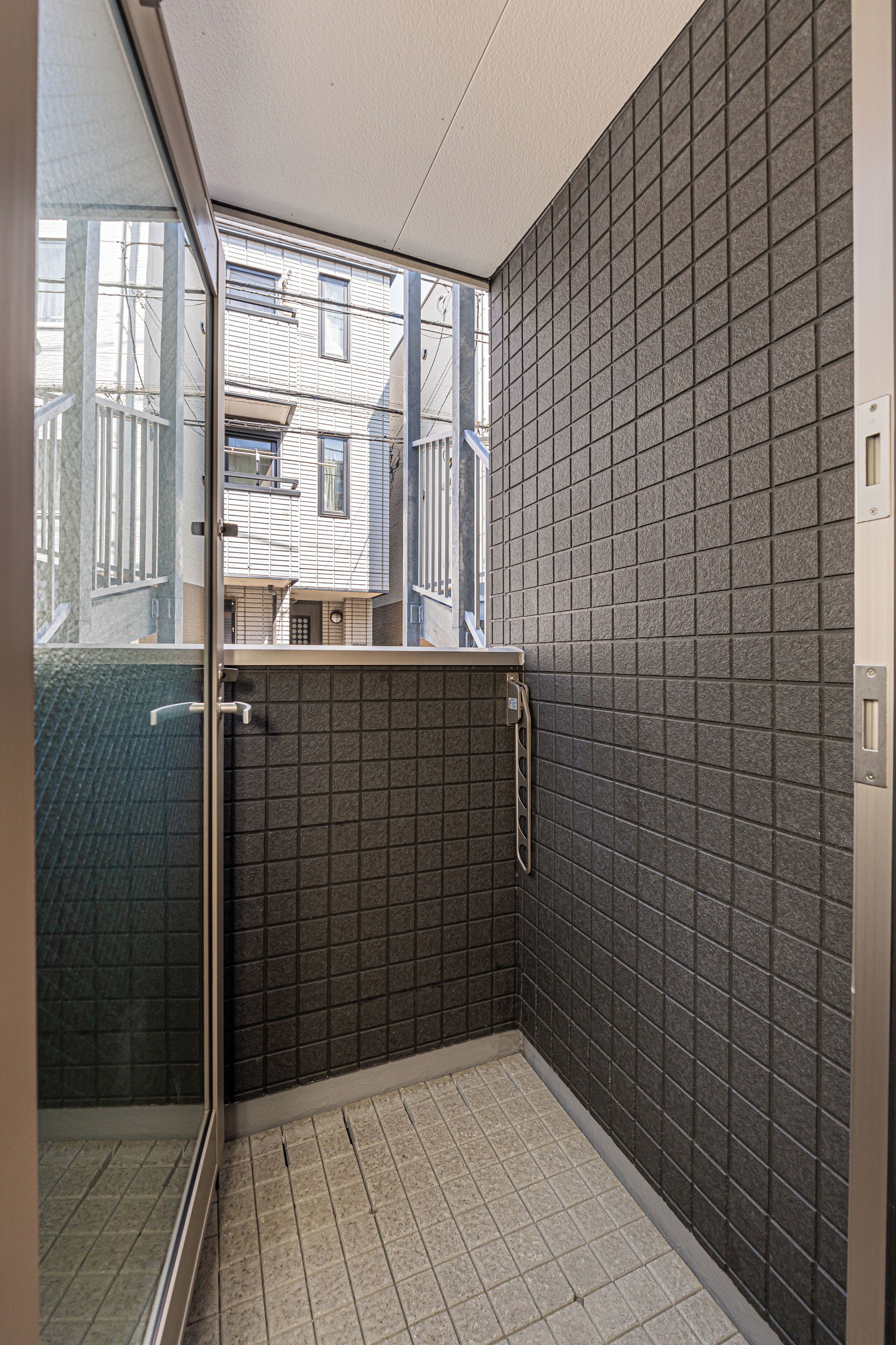 A small, enclosed bathroom with a tiled floor and walls, a glass door leading outside, and a window providing a view of the outside.