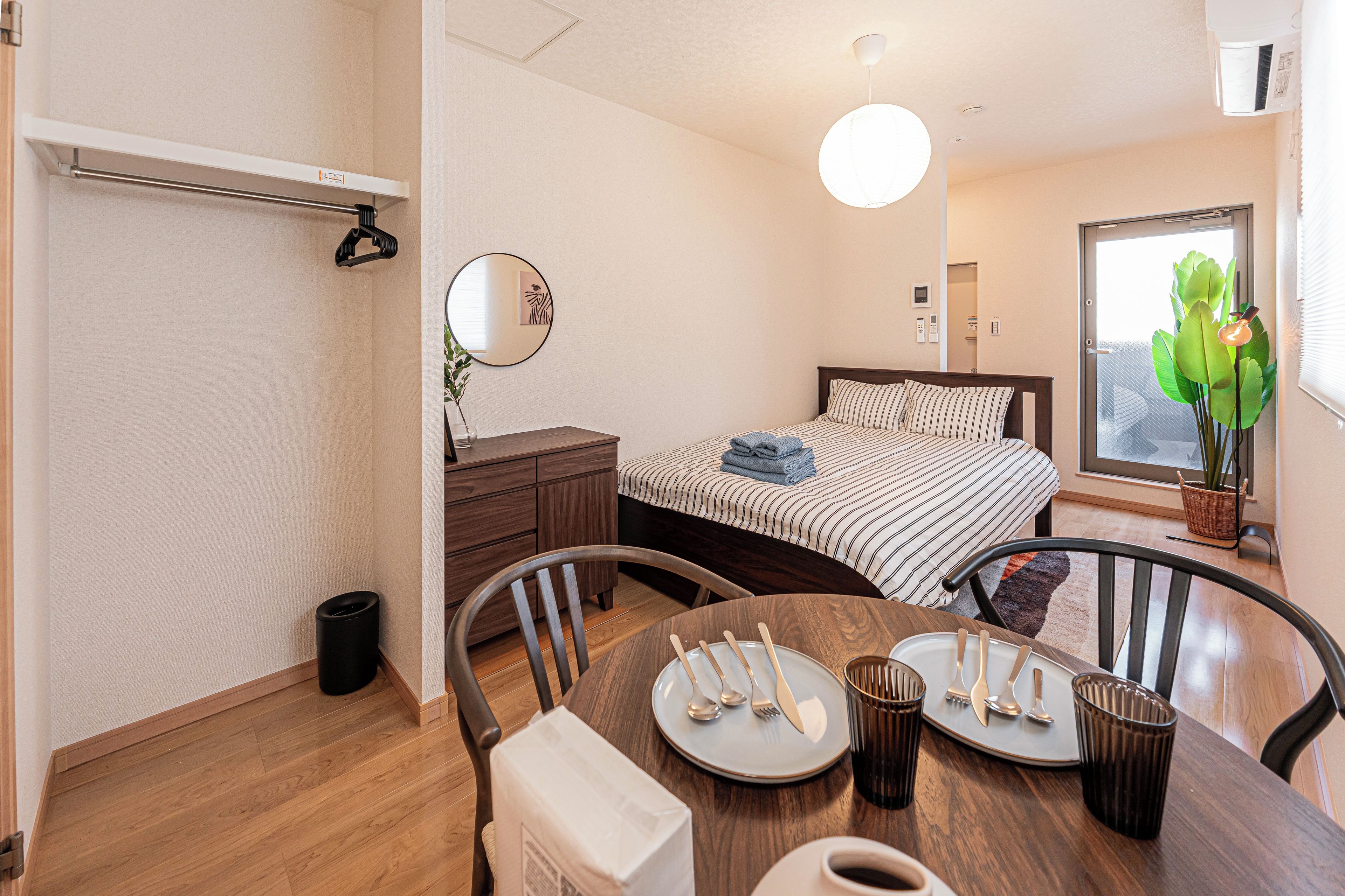 Habitación de hotel con cama, mesita de noche, silla, mesa de comedor con cubiertos, un cubo de basura, lámpara colgante, espejo en la pared y ventana con vista a follaje.