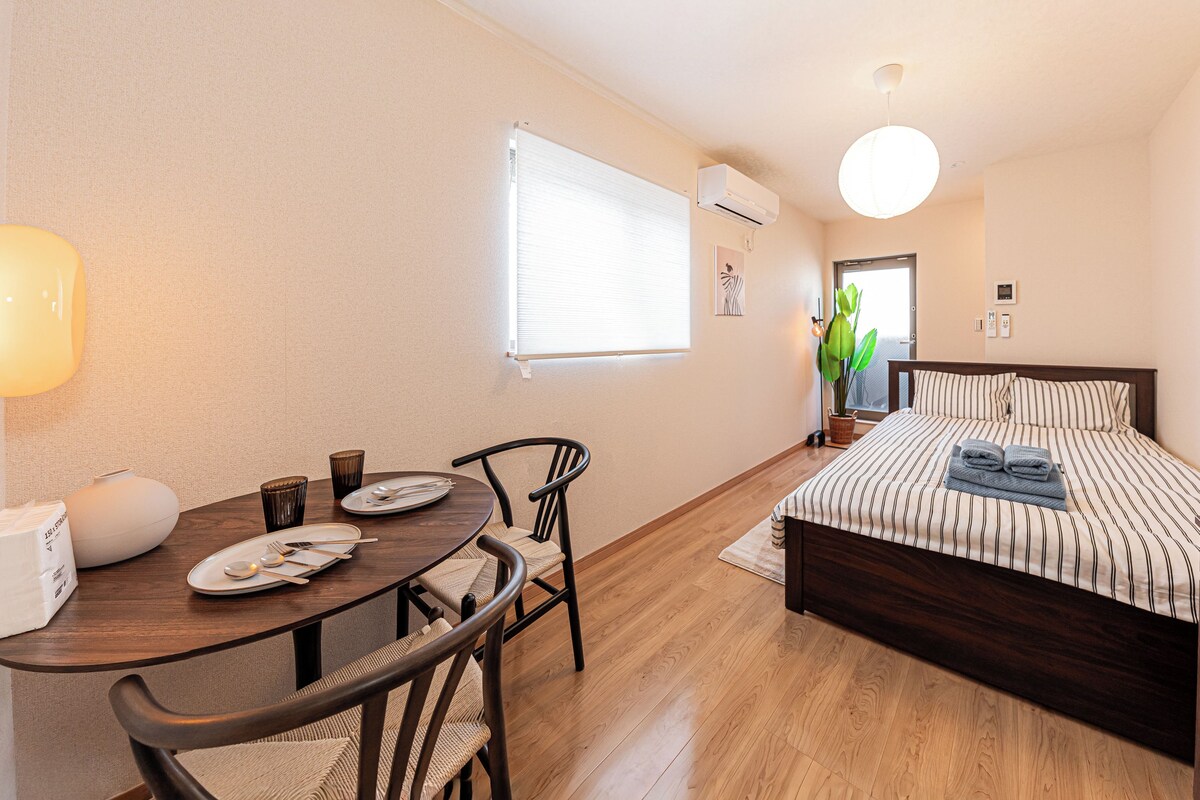 A hotel room with a wooden floor, a bed with striped bedding, a dining table with chairs, and a wall-mounted air conditioner.