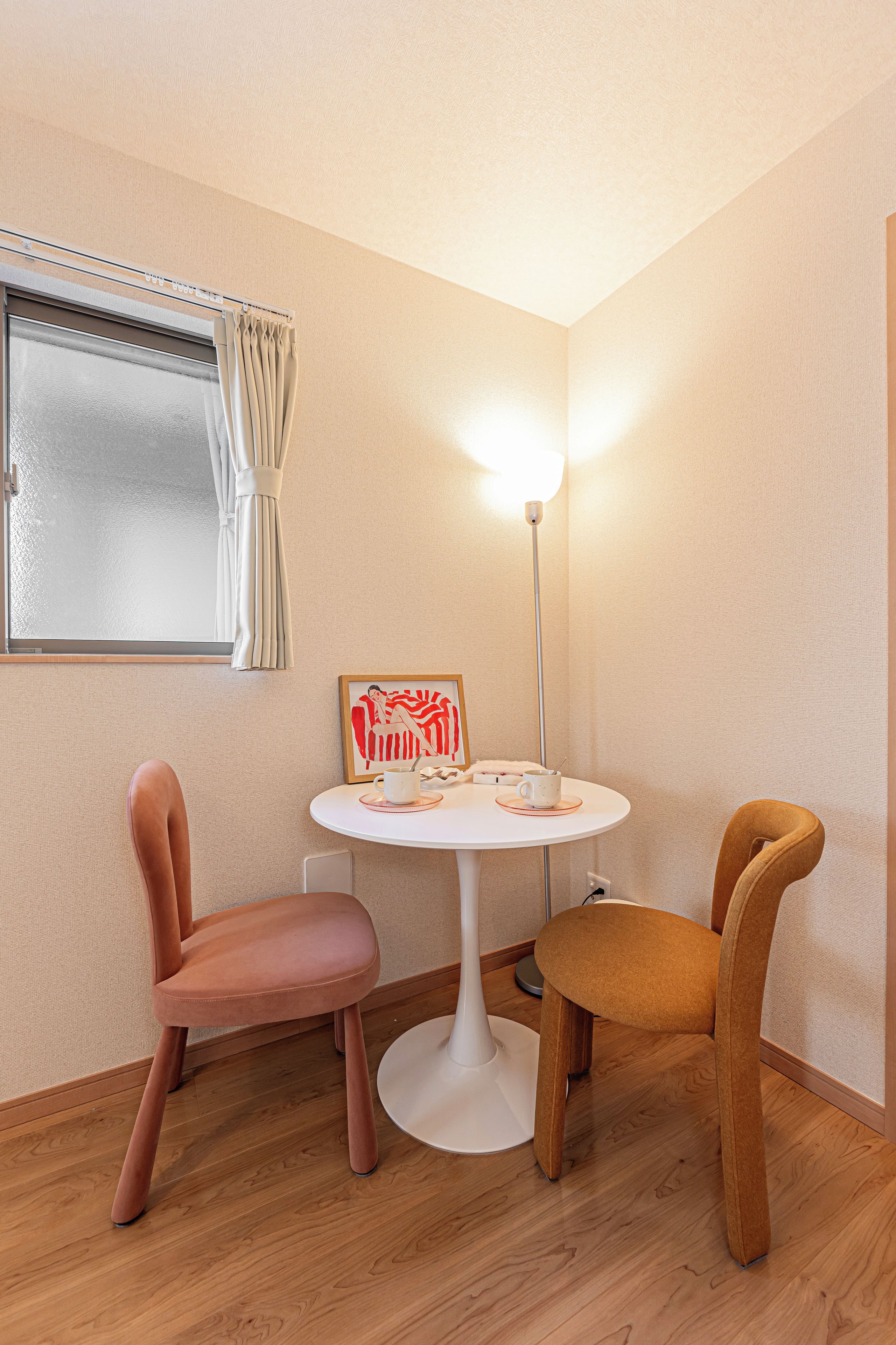 A corner room with a round table, two chairs, a window with curtains, a floor lamp, and a framed picture on the wall.