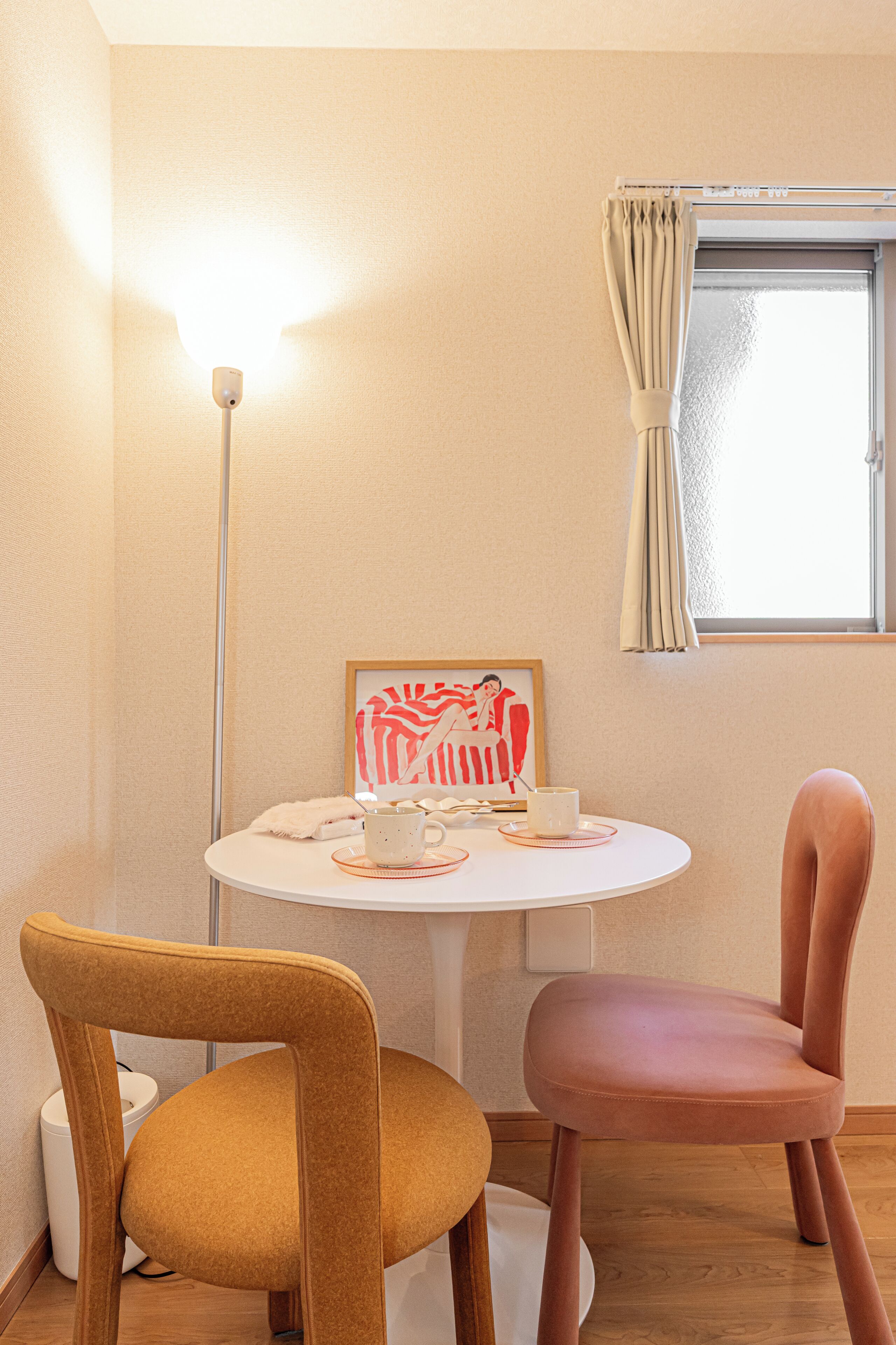 Une petite pièce avec une table ronde, deux chaises, un lampadaire, une fenêtre avec des rideaux et une œuvre d’art encadrée accrochée au mur.