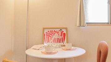 Une petite pièce avec une table ronde, deux chaises, un lampadaire, une fenêtre avec des rideaux et une œuvre d’art encadrée accrochée au mur.