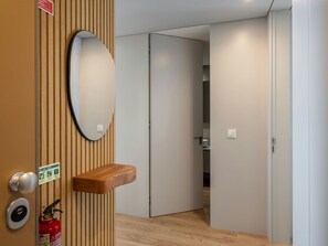 A hallway with a wooden panel wall, a round mirror, a wooden shelf, a fire extinguisher, and a door leading to a bathroom. - Azure Calheta by Interhome (Calheta (Madeira))