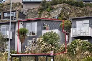 A modern house with a balcony, surrounded by vegetation and a rocky hillside. - Spacious 3 BDRM, Panoramic Views and 2 Carparks! (Queenstown)