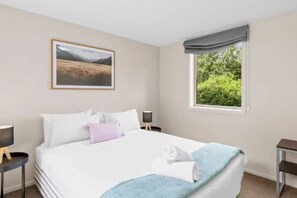 A hotel room with a large bed, a window with a view of greenery, a framed picture on the wall, and a bedside table with a lamp. - Spacious 3 BDRM, Panoramic Views and 2 Carparks! (Queenstown)