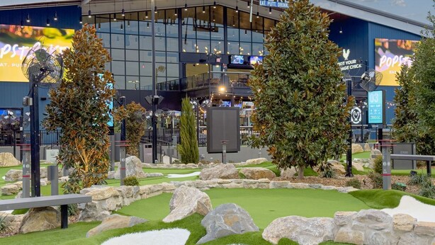 Un giardino interno con erba sintetica, rocce e alberi, circondato da un edificio con ampie finestre e schermi digitali.