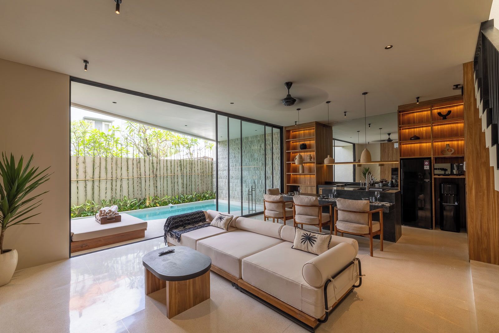 A modern living room with a sofa, coffee table, and dining area. A kitchen is visible in the background with a dining table and chairs.
