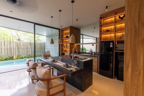 A modern kitchen with a central island, bar seating, and a view of a swimming pool. - Sila Villa Berawa - By Saamu (Badung)