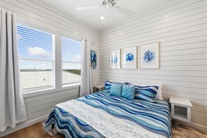 A bedroom with a bed, a nightstand, a ceiling fan, and a view of the beach through the window.