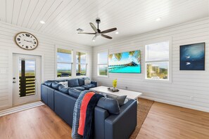 A modern living room with a sectional sofa, a ceiling fan, and large windows with views of the outdoors.