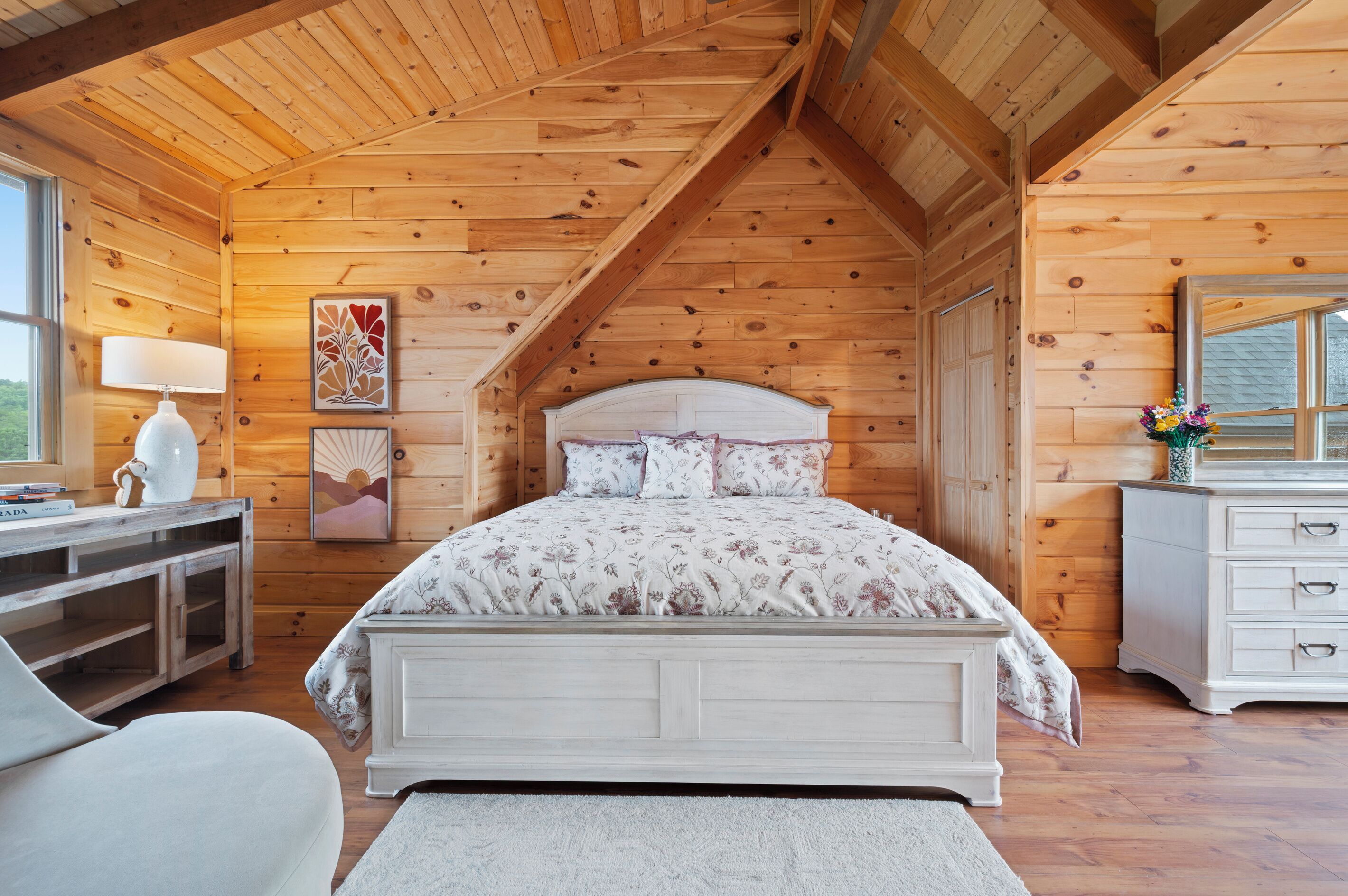 Une chambre avec un plafond en bois, un lit recouvert d’une literie à motifs floraux, un bureau avec une lampe et une commode avec un miroir.