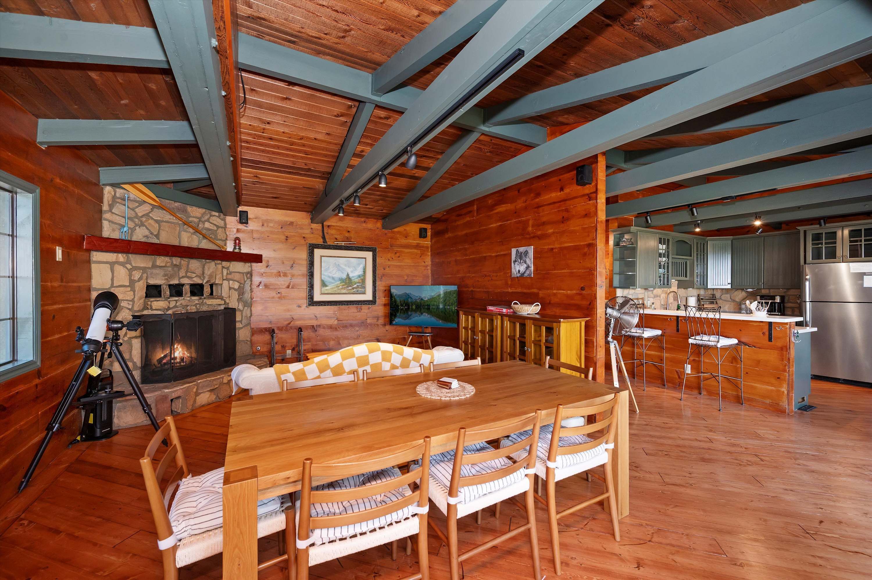 A cozy cabin interior with a stone fireplace, wooden dining table, and kitchen area.