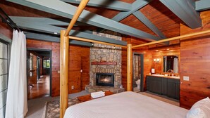Ein Blockhaus-Interieur mit Kamin aus Naturstein, Holzbalken, einem Bett und einem Badezimmer mit Waschtisch und Spiegel.