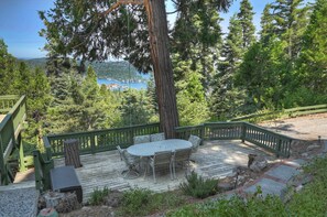 A wooden deck with a round table and chairs, surrounded by a green railing, overlooking a lake and forested area.