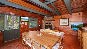 Interior de una cabaña con comedor, chimenea, televisor y un cartel que indica 'CAMP HOST'.