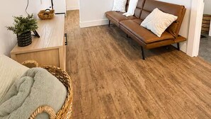 A modern living room with a brown leather sofa, a flat-screen TV mounted on the wall, a wooden cabinet, and a wicker basket. - Harbour Suite, Modern Stay Near NFLD ferry (North Sydney)