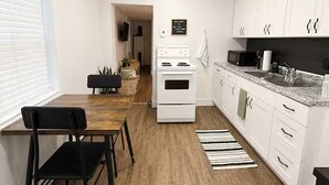 A modern kitchen with white cabinets, a stove, and a dining area with a black chair and table. - Harbour Suite, Modern Stay Near NFLD ferry (North Sydney)