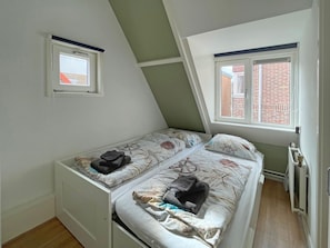 A small, single-room apartment with a sloped ceiling, two beds with patterned bedding, a radiator, and two windows with views of a brick building. - SunnyLogde by Interhome (Noordwijk)