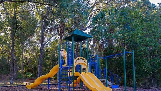 Ein Spielplatz mit zwei Rutschen und einer Kletterkonstruktion, umgeben von Bäumen.