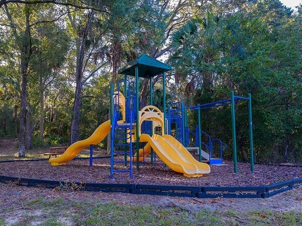 Ein Spielplatz mit zwei Rutschen und einer Kletterkonstruktion, umgeben von Bäumen.
