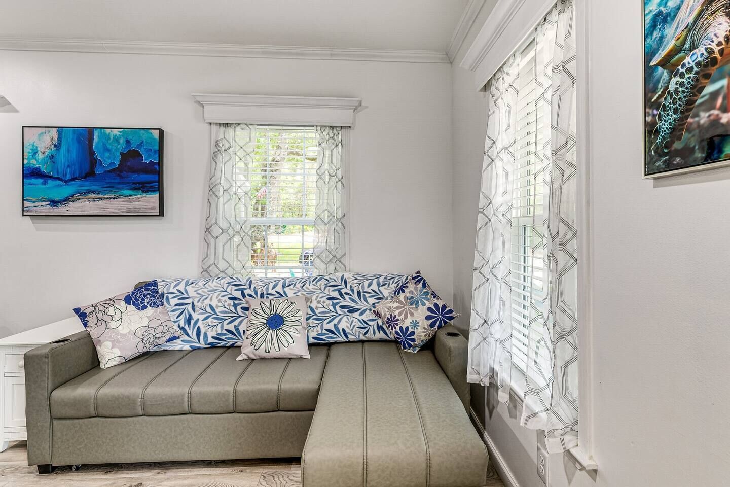 A room with a grey sofa, patterned cushions, a painting on the wall, and a window with patterned curtains.