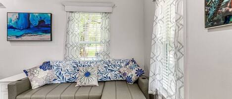 A room with a grey sofa, patterned cushions, a painting on the wall, and a window with patterned curtains.
