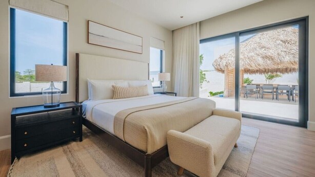 Een moderne slaapkamer met een groot bed, nachtkastjes, een bankje en uitzicht op het strand door grote ramen.