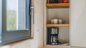 Een kitchenette met een koffiezetapparaat, een plank met bekers en een raam.