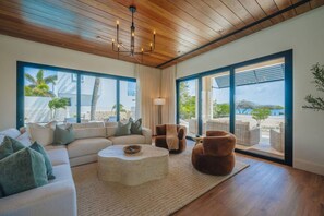 A modern living room with a wooden ceiling, large windows, a sofa, armchairs, and a coffee table. - New Oceanfront LUX 5BR Pool · Sunset Cove Malmok (Noord)