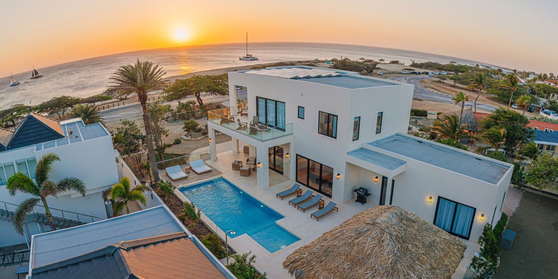 A modern two-story house with a swimming pool, large windows, and a view of the ocean at sunset.