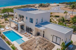 A modern two-story house with a swimming pool, large windows, and a view of the beach. - New Oceanfront LUX 5BR Pool · Sunset Cove Malmok (Noord)