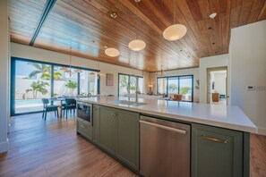 A modern kitchen with a large island, wooden ceiling, and large windows overlooking a pool area. - New Oceanfront LUX 5BR Pool · Sunset Cove Malmok (Noord)