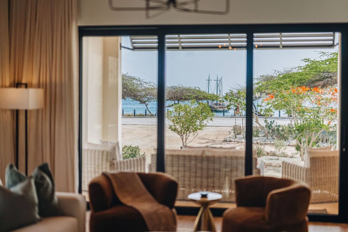 Quarto com grande porta de correr de vidro que dá para uma vista da praia, móveis de vime e uma luminária.