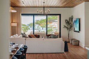 Ein modernes Wohnzimmer mit einem Ecksofa, einem Essbereich mit Tisch und Stühlen, einem großen Fenster mit Aussicht und einer Hängelampe.