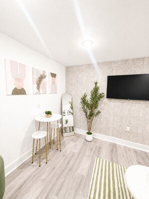 Uma sala de estar moderna com uma TV de tela plana, uma mesa de jantar redonda com banquetas, um vaso de plantas e arte abstrata na parede.