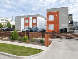 Ein modernes Wohngebäude mit grau-roter Fassade, ein Parkplatz mit mehreren Autos und ein gepflegter Garten mit Pflanzen und einem Holzzaun.
